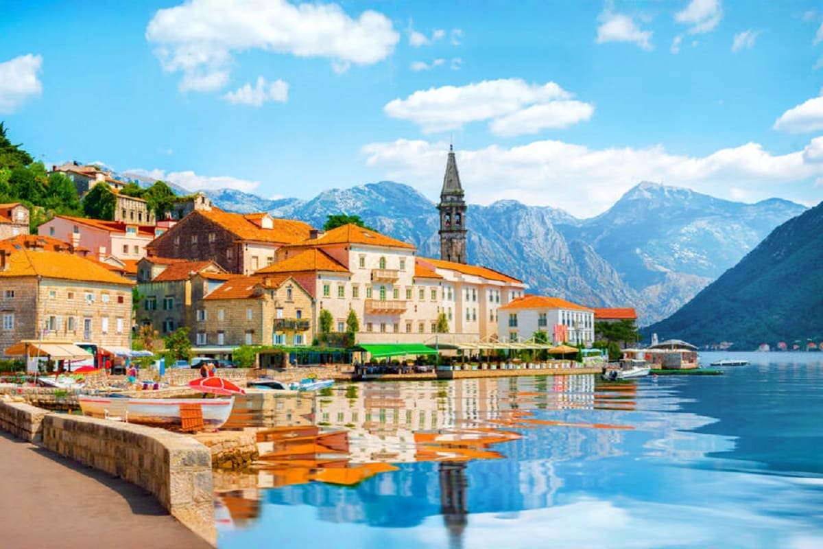 Picturesque coastal town with colorful buildings, orange roofs, and church bell tower reflected in calm blue bay waters, surr