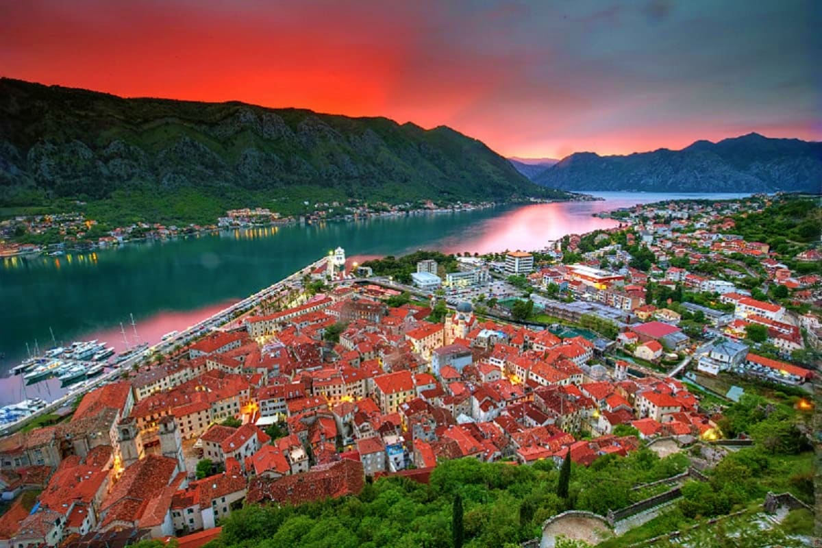 Aerial sunset view of Kotor Old Town with red-roofed buildings, marina, and surrounding mountains reflected in blue bay water