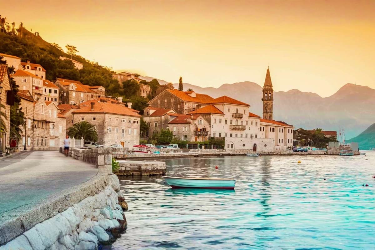 Coastal town at sunset with stone buildings, terracotta roofs, church bell tower, and turquoise Mediterranean waters with fis