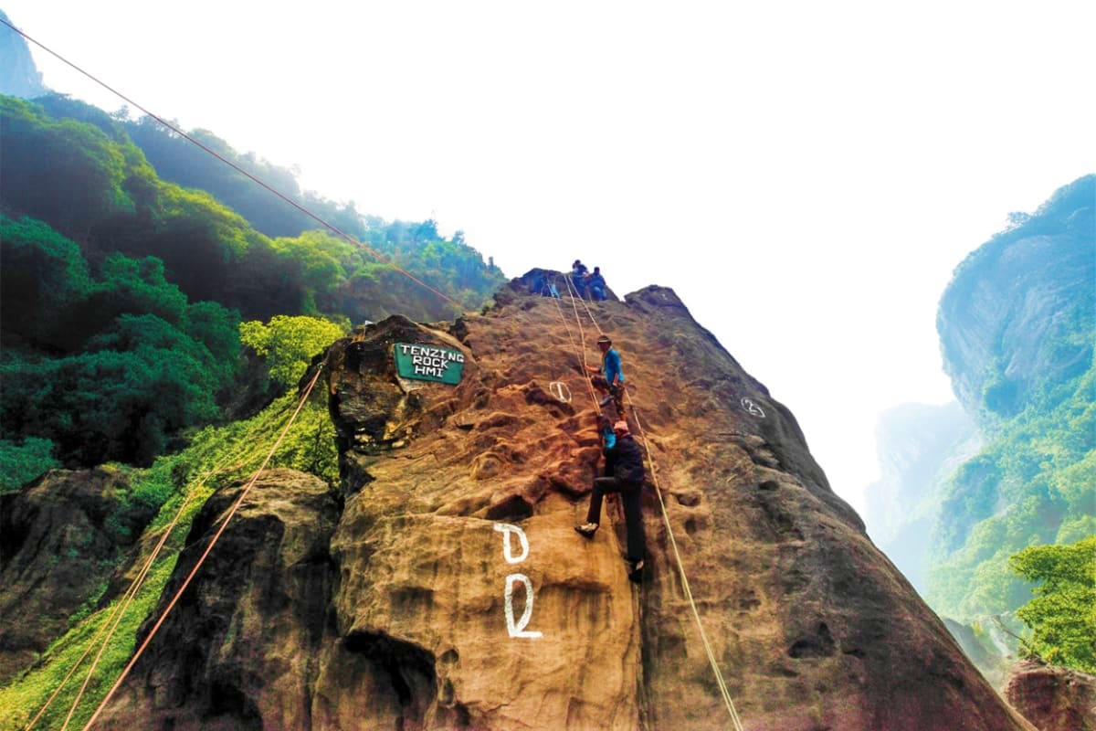 Rock climbers rappelling down a steep cliff face marked with "DQ" in a lush mountainous valley with forested peaks