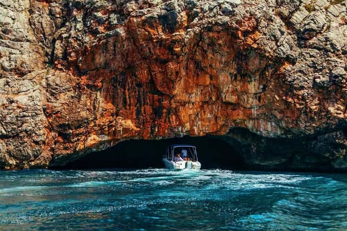 Small white boat entering a sea cave beneath russet and gray rocky cliffs in turquoise Mediterranean waters
