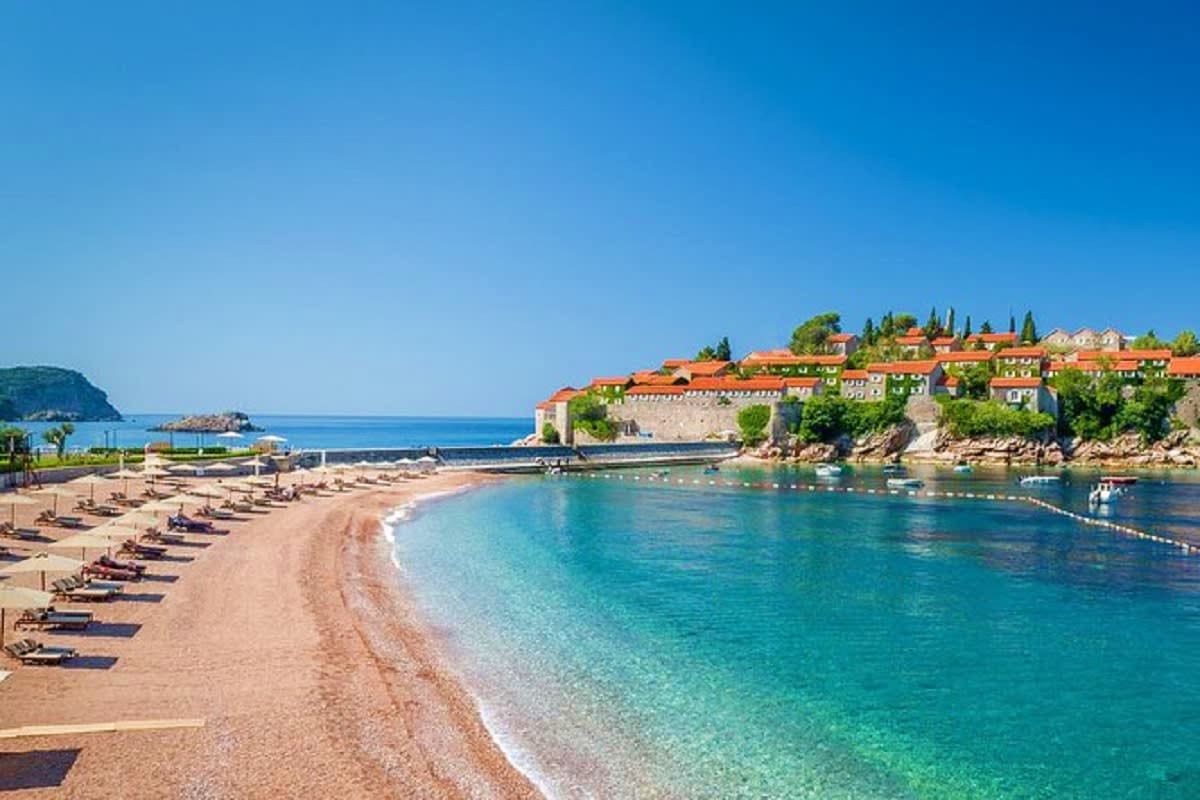 Sveti Stefan island beach with turquoise waters, sandy shore lined with lounge chairs, and terracotta-roofed buildings on roc