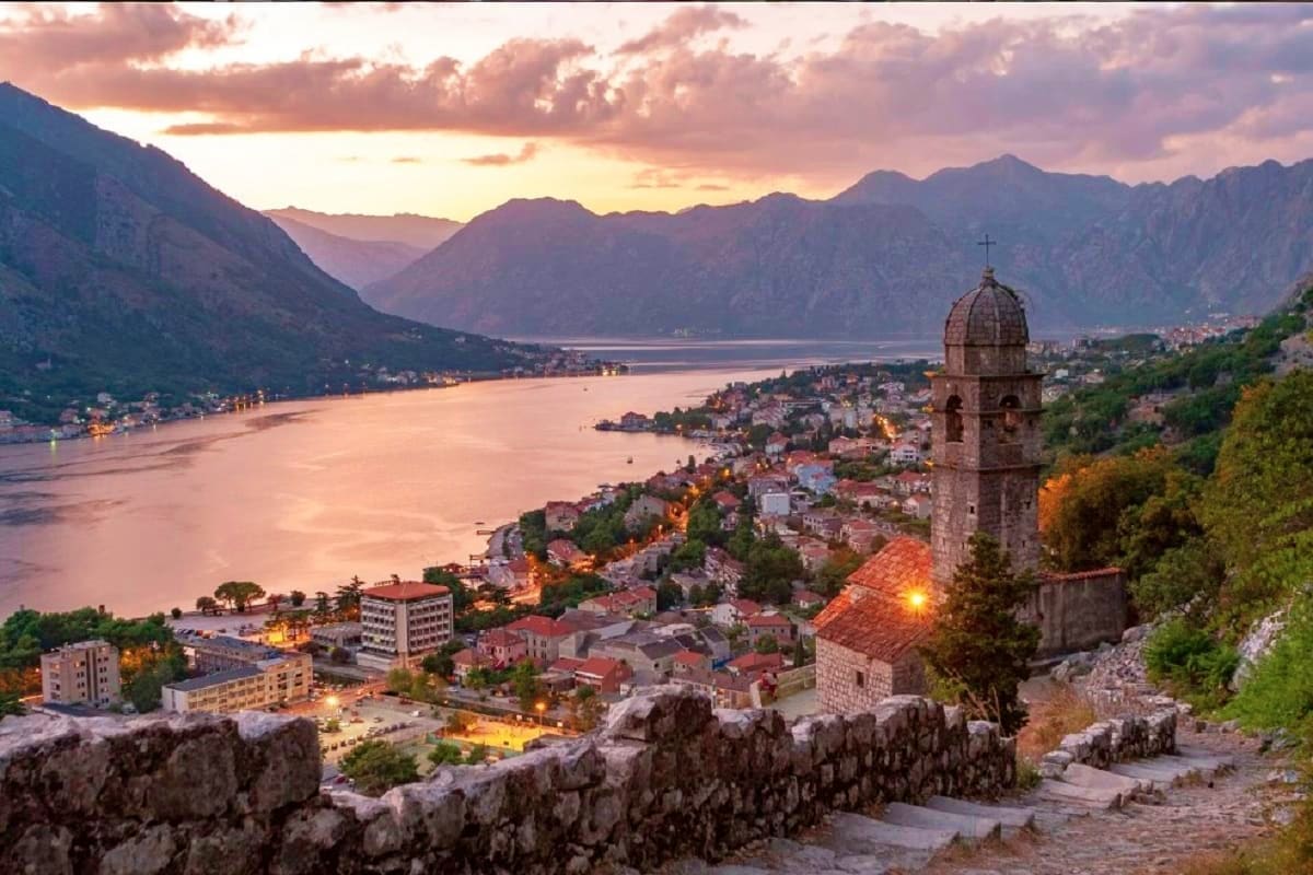 Kotor Bay at sunset with a stone church bell tower in the foreground, surrounded by mountains and a waterfront town with glow
