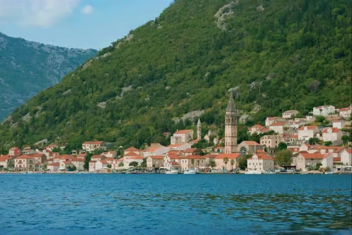Coastal town with terracotta-roofed buildings, church towers, and forested mountains rising behind, viewed from blue water