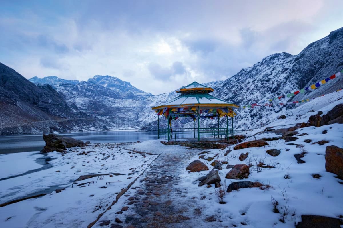Snow-covered lakeside gazebo with colorful prayer flags at Gurudongmar Lake, surrounded by snow-capped mountains in Sikkim