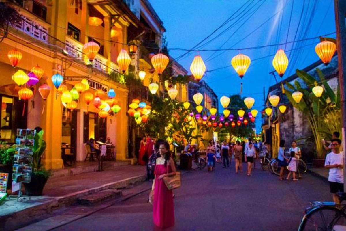 Hoi An ancient town street at dusk with colorful silk lanterns hanging overhead, yellow buildings, and pedestrians walking th