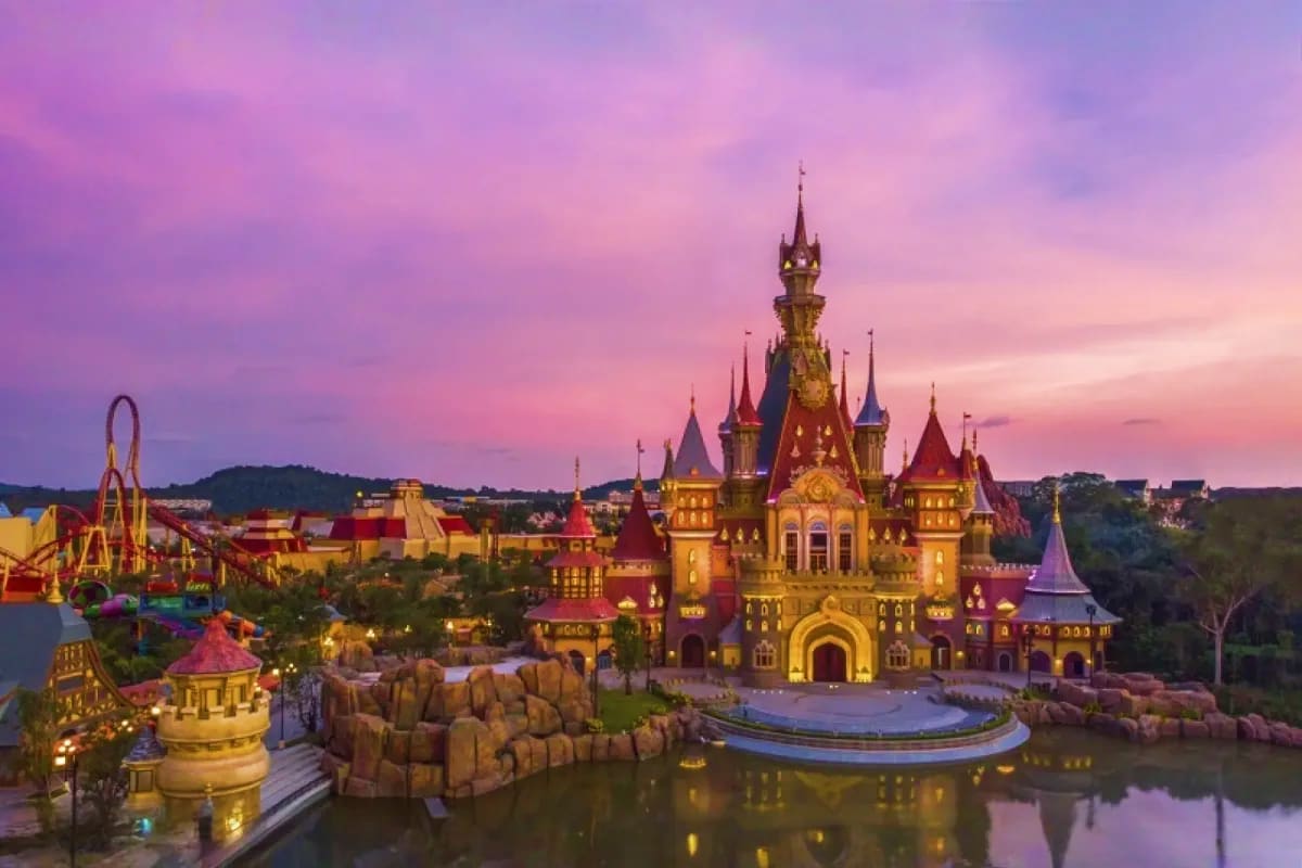 Illuminated fantasy castle at dusk with purple and pink sky, surrounded by theme park attractions and water reflection in Vie