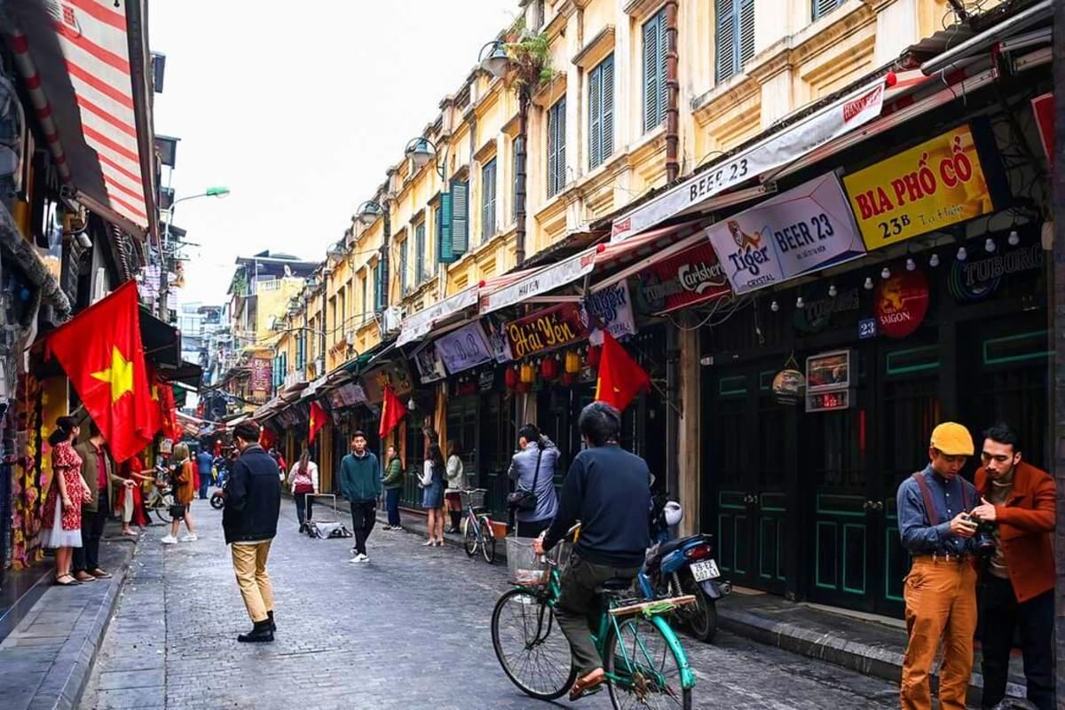 Busy street in Hanoi's Old Quarter with French colonial architecture, Vietnamese flags, restaurants, cyclists, and pedestrian