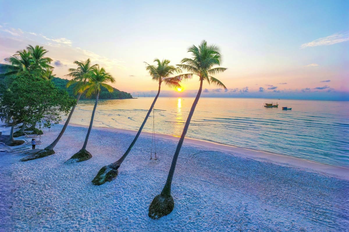 Tropical beach at sunset with palm trees leaning over white sand, calm turquoise water, and fishing boats, Vietnam