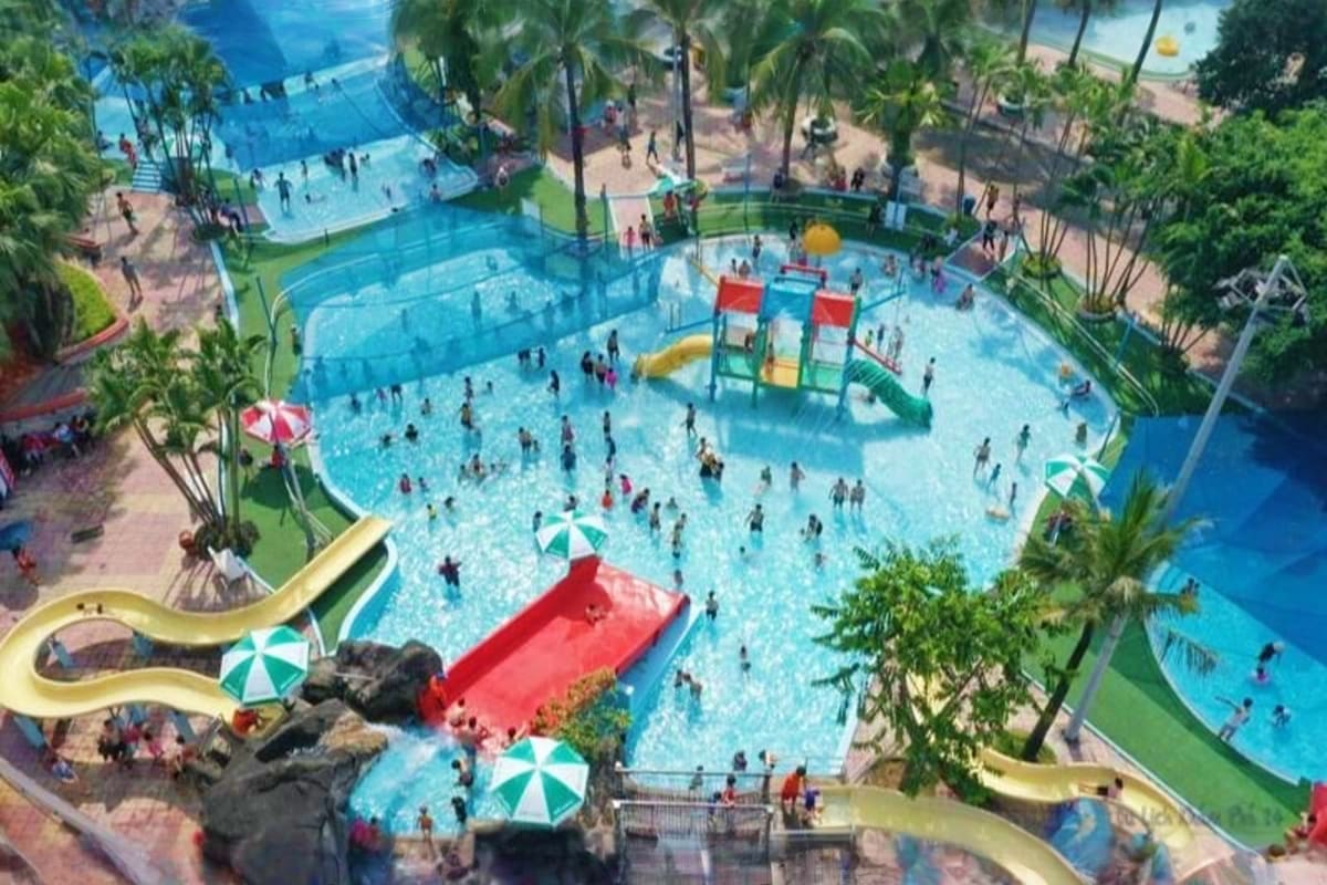 Aerial view of a busy water park with multiple swimming pools, colorful water slides, palm trees, and visitors enjoying vario