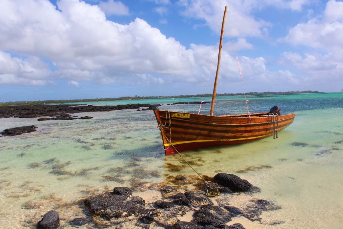 Traditional wooden fishing boat anchored in turquoise shallow waters off a Mauritius beach with black rocks and white sand.
