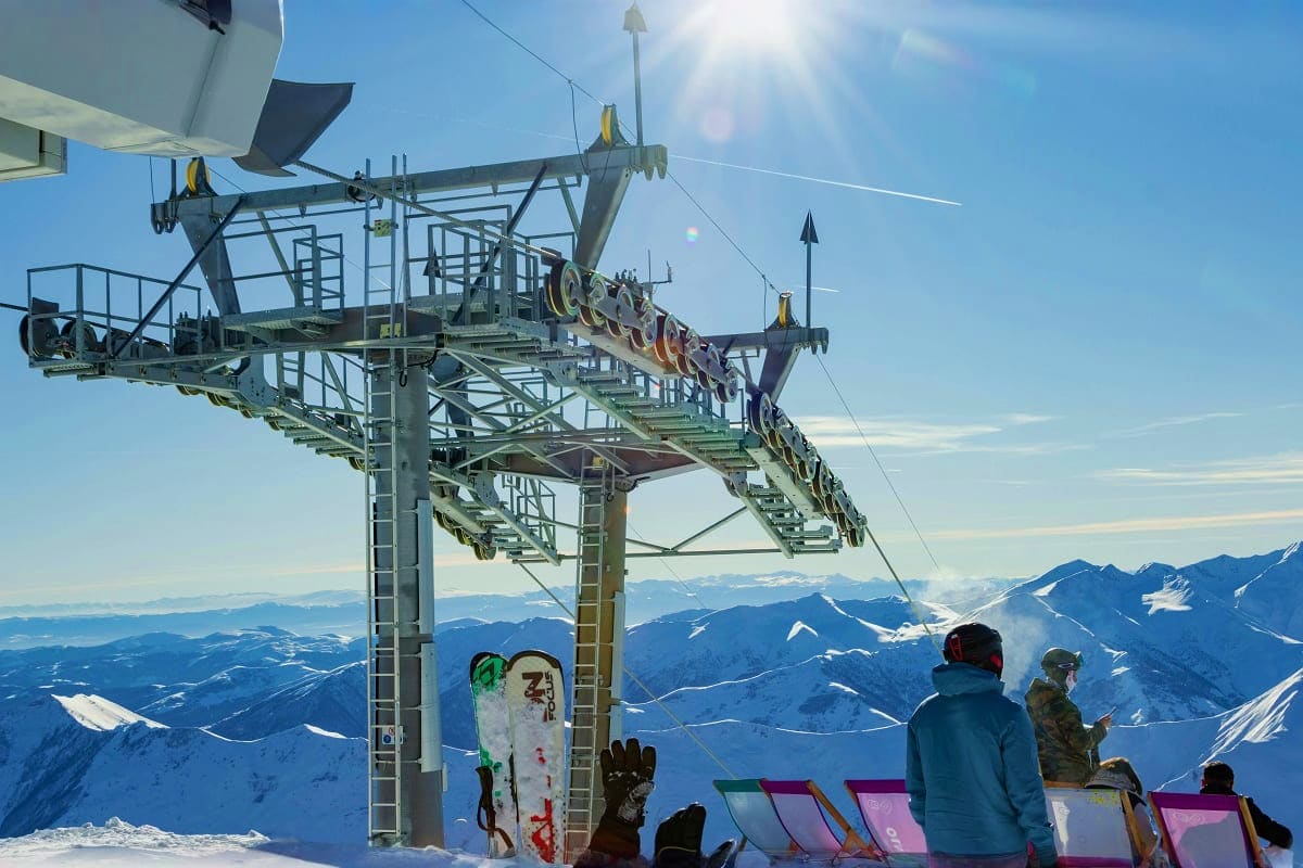 Snowboarders and skiers at mountain ski lift with snowy Alpine peaks and valley landscape in background under blue sky