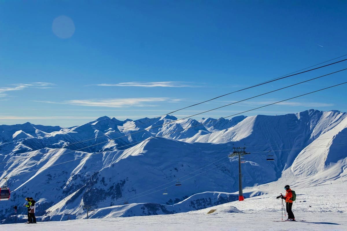 Skier on snowy mountain slope with ski lift cables and snow-covered peaks in background under clear blue sky