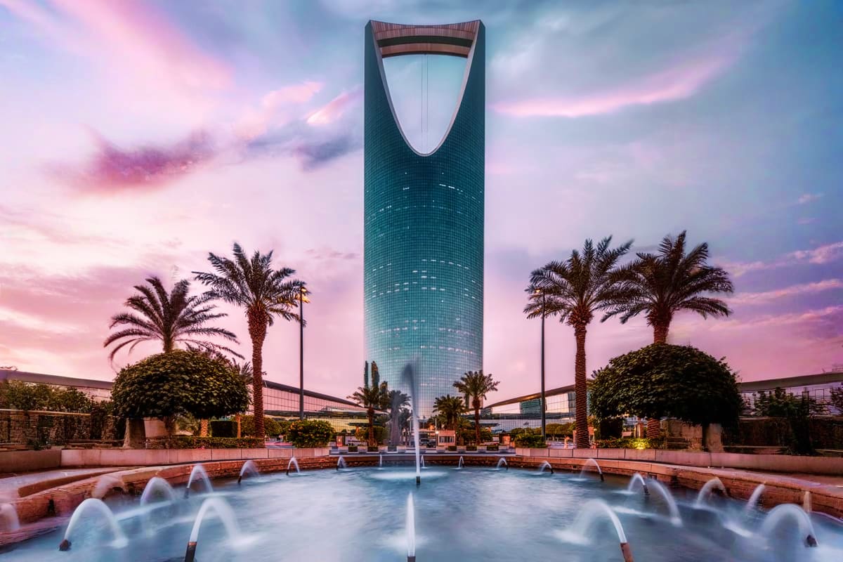 Kingdom Centre tower in Riyadh at sunset with illuminated fountains, palm trees, and pink sky