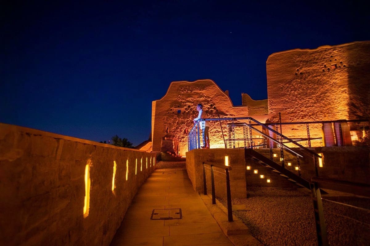 Ancient mud brick fortress ruins illuminated at night with golden lighting and walkway under deep blue sky in Riyadh