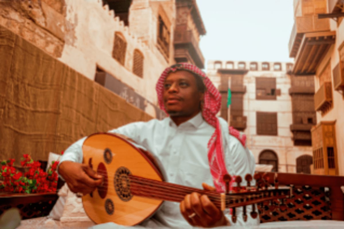Man wearing white shirt and pink ghutra playing oud in historic Jeddah courtyard with traditional architecture and red flower