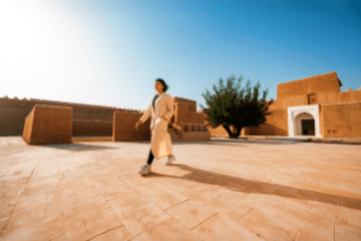Woman walking through historic clay-brick courtyard with traditional architecture under blue sky in Jeddah