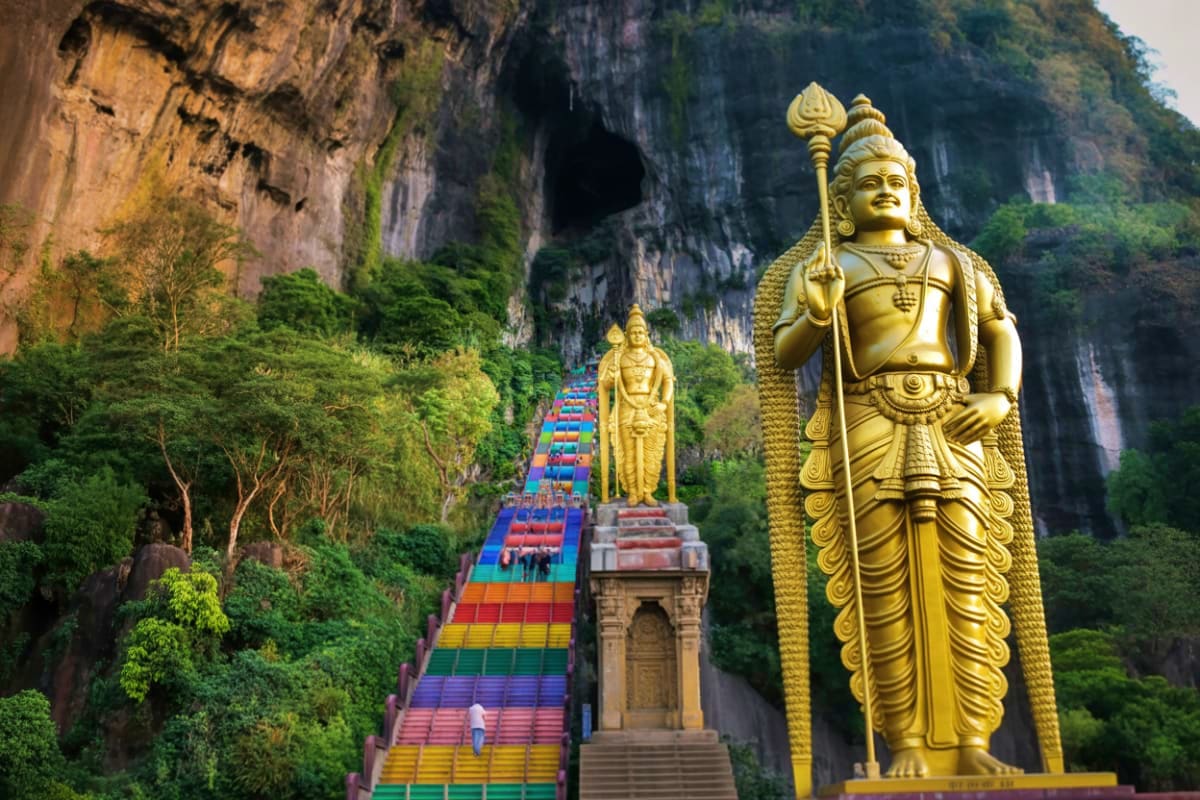 Singapore Malaysia Family tour Golden Hindu deity statue at Batu Caves entrance with colorful rainbow staircase and limestone cliff face in Malaysia