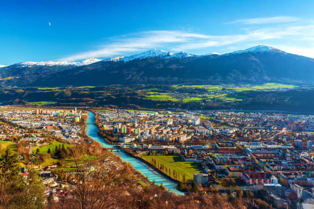 Central Europe Romance Tour With Prague to Venice for Couples Aerial view of Innsbruck Austria with turquoise river winding through city, snow-capped Alps mountains in background under bl
