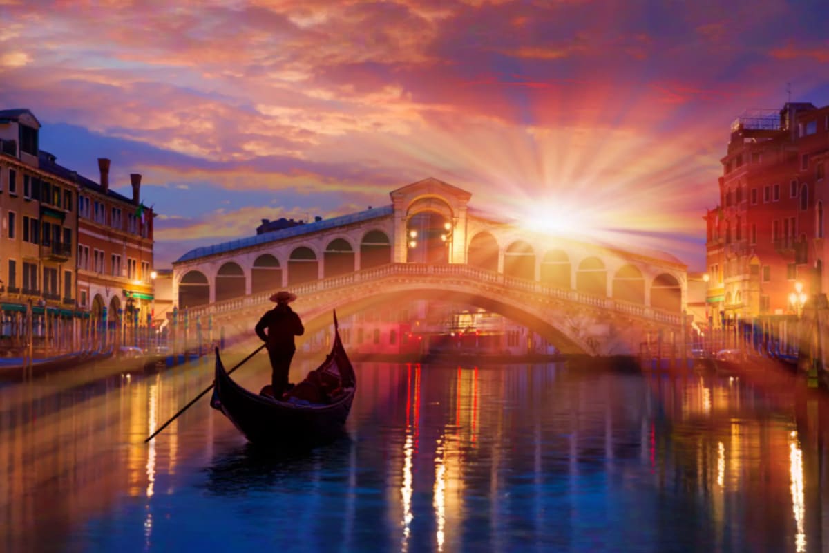 Gondolier punting a gondola on Venice's Grand Canal at sunset with the Rialto Bridge illuminated in the background