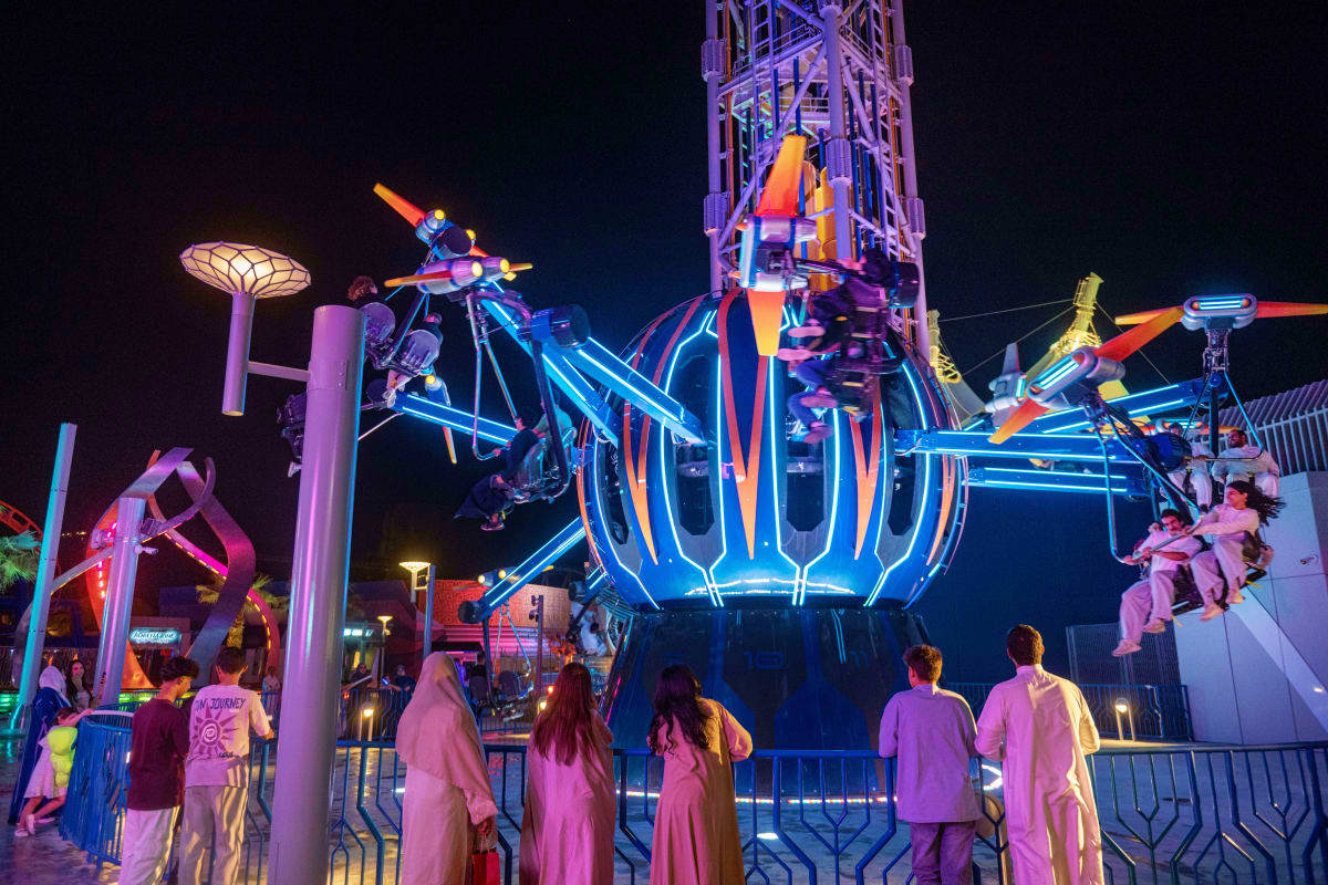 Nighttime amusement park ride with bright neon blue and orange lighting, visitors watching from barrier below, tall illuminat