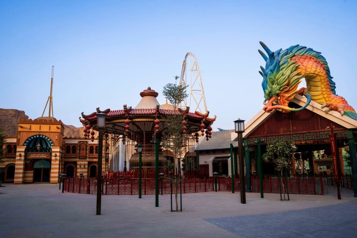 Amusement park plaza with colorful dragon statue, Chinese-style pavilion, roller coaster track, and ornate red-fenced structu