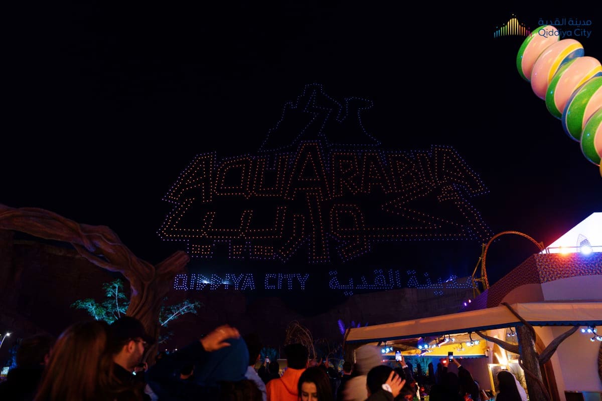 Nighttime drone light show spelling "Qiddiya City" above a crowd at an adventure theme park with illuminated rides and struct