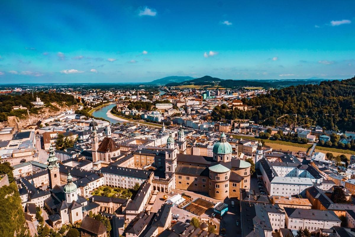 Aerial view of Salzburg, Austria with cathedral domes, Salzach River, surrounding mountains and forested hills under blue sky