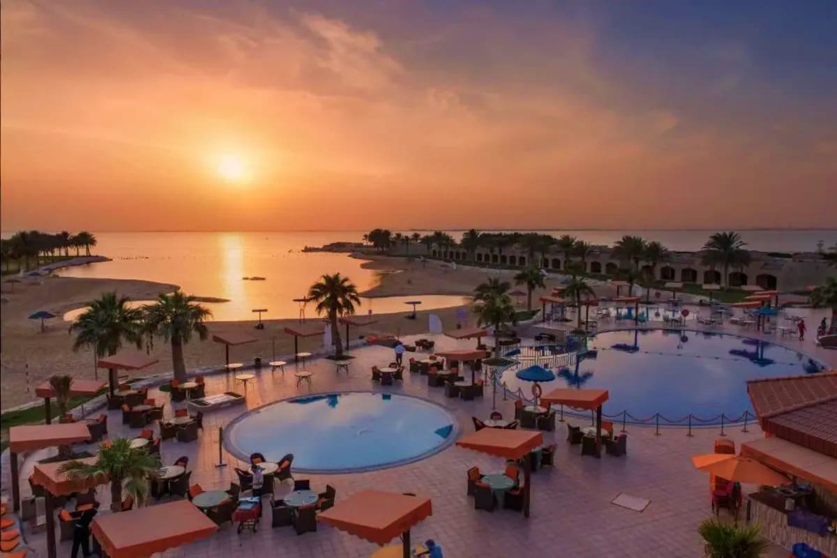 Aerial view of a beachfront resort in Dammam at sunset with pools, palm trees, sandy beach, and calm waters