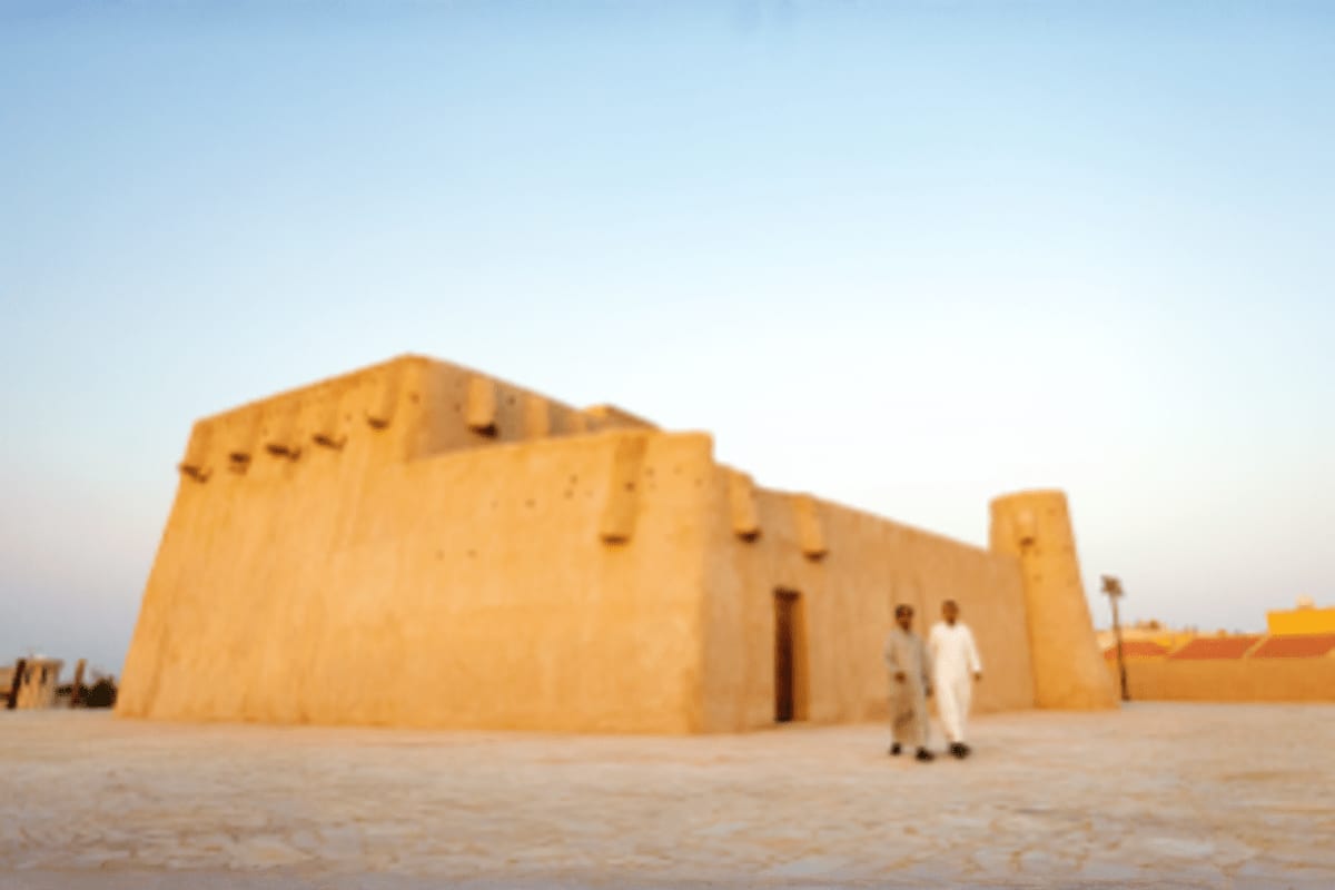 Adobe mud brick fortress with two people in white traditional clothing standing in desert landscape under clear sky