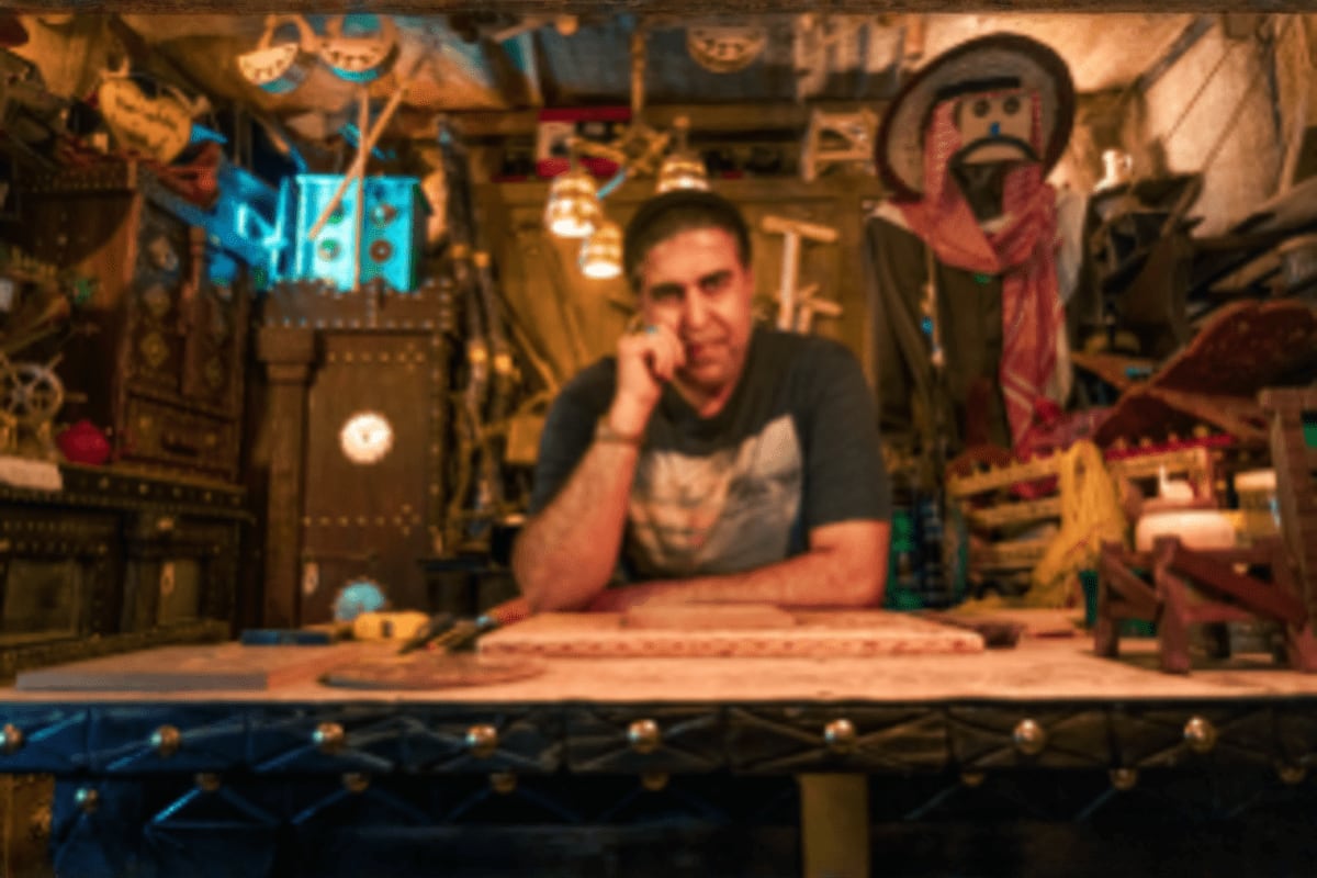 Artist in steampunk workshop surrounded by mechanical sculptures and neon signs, leaning on workbench with tools