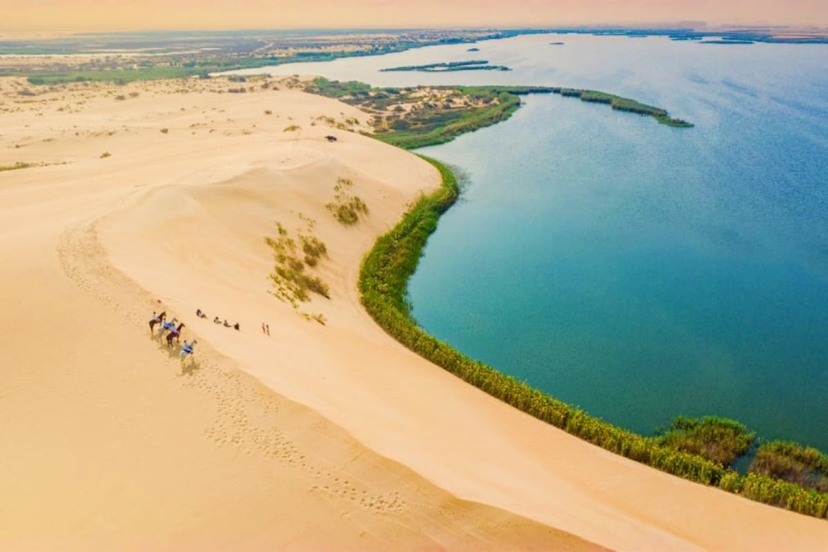 Aerial view of sand dunes meeting turquoise water with green vegetation and visitors on the beach exploring the coastal lands