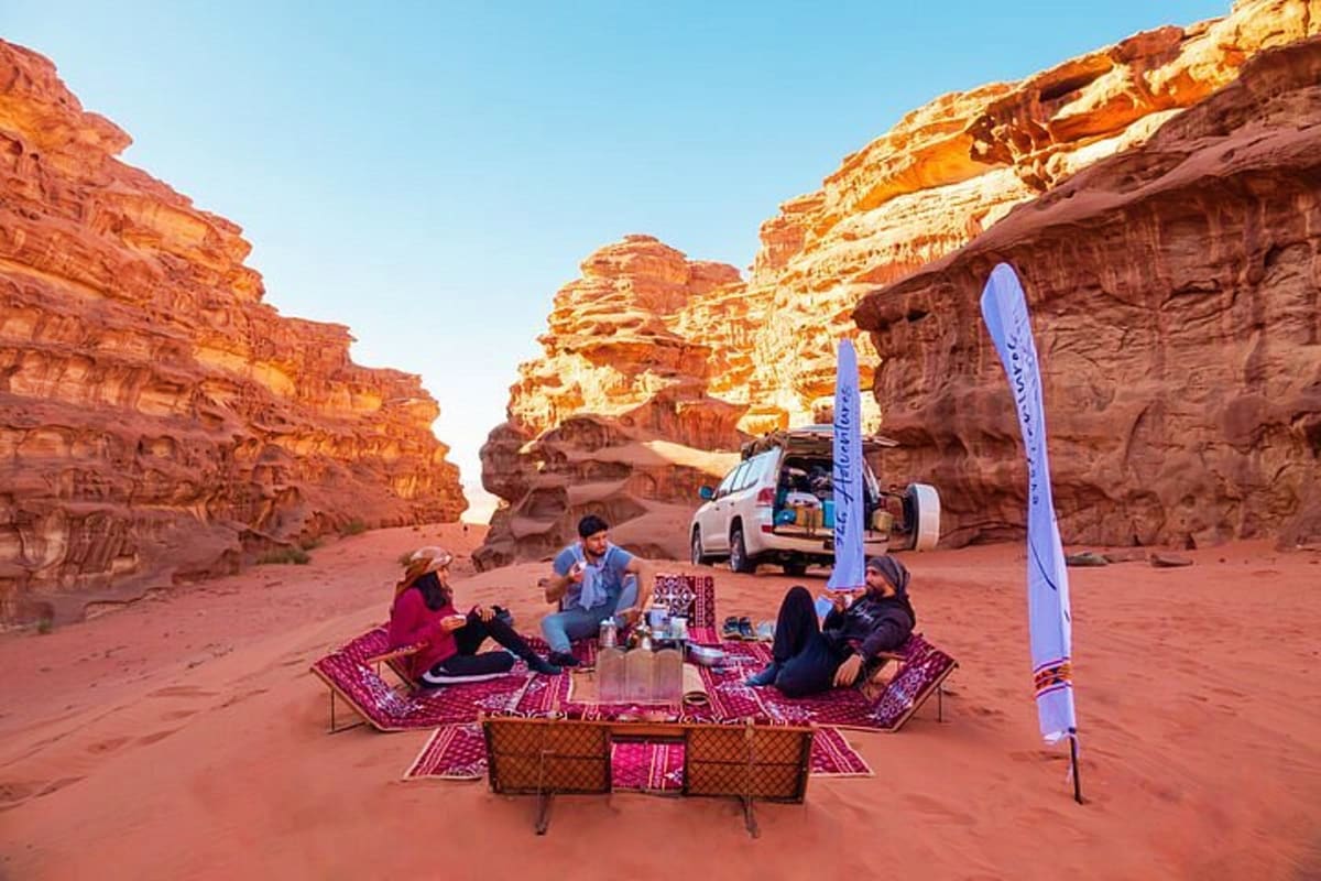 Group camping in a red desert canyon with towering sandstone cliffs, traditional rugs, and a vehicle parked nearby under clea