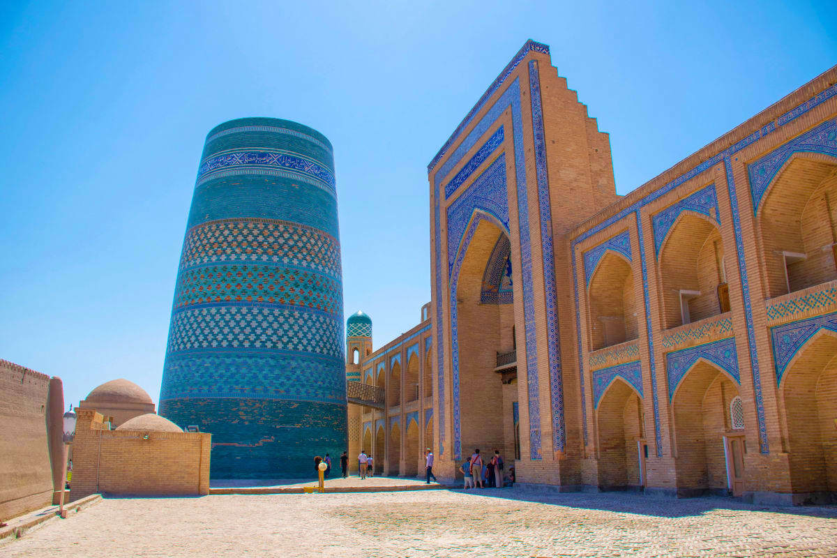 Historic Islamic architecture in Khiva with turquoise minaret and ornate blue tilework on courtyard buildings under clear sky