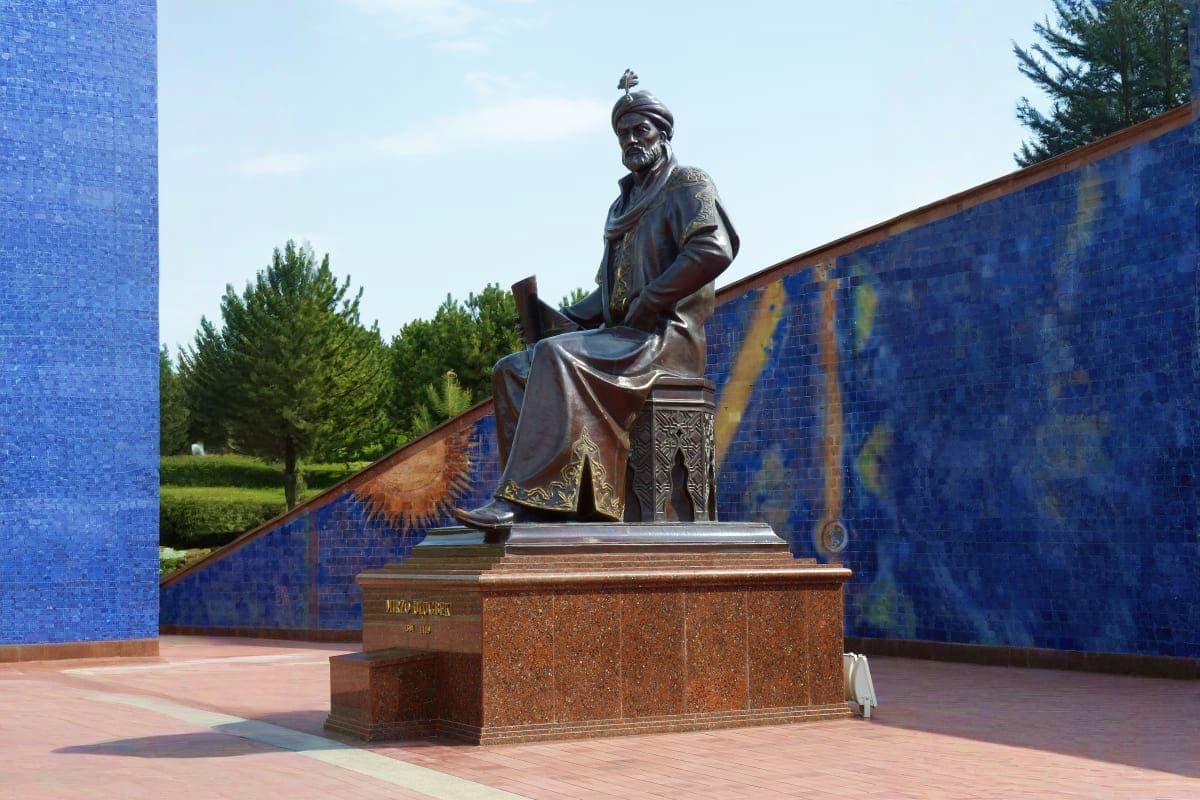 Bronze statue of a robed historical figure wearing a turban, seated on ornate pedestal in plaza with blue tiled walls and gre