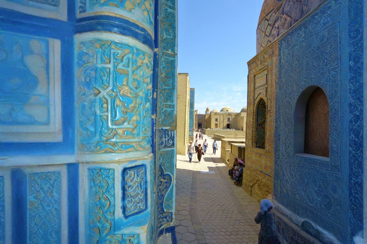 Blue and gold ornamental tilework frames a narrow courtyard passage with Islamic architecture, visitors walking through an an