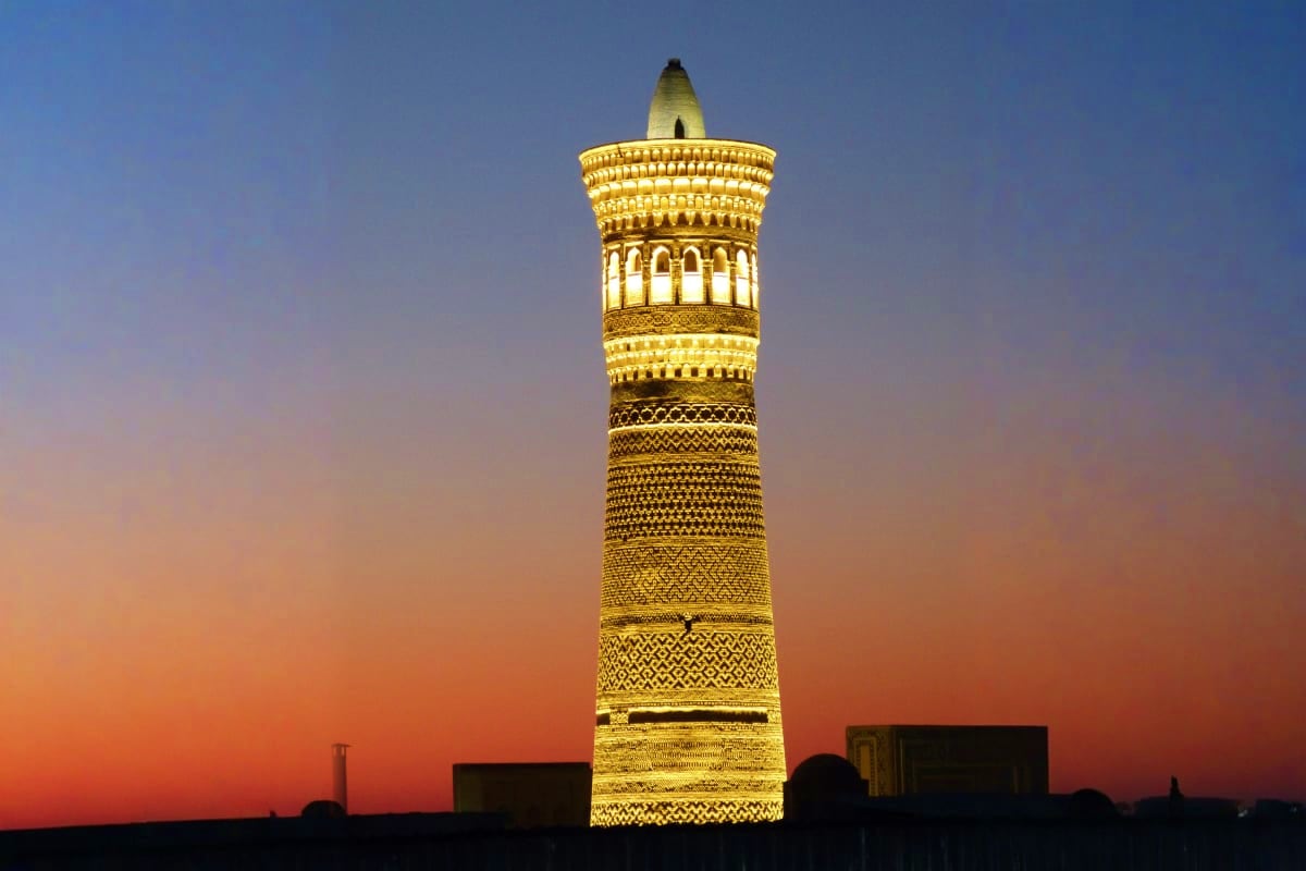 Illuminated minaret tower with ornate brick patterns and arched details glowing at dusk against a gradient sky in Uzbekistan