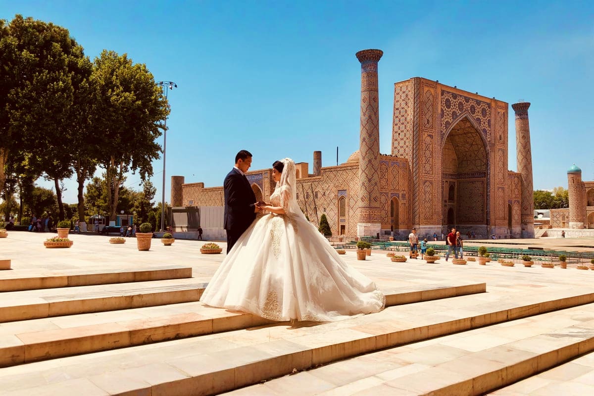 Bride and groom in traditional wedding attire at Registan Square in Samarkand, Uzbekistan, with ornate Islamic architecture a