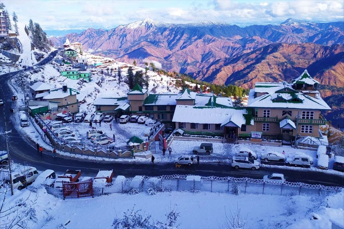 Snowy mountain resort buildings with snow-covered Himalayan peaks and forested valleys in background at Manali hill station