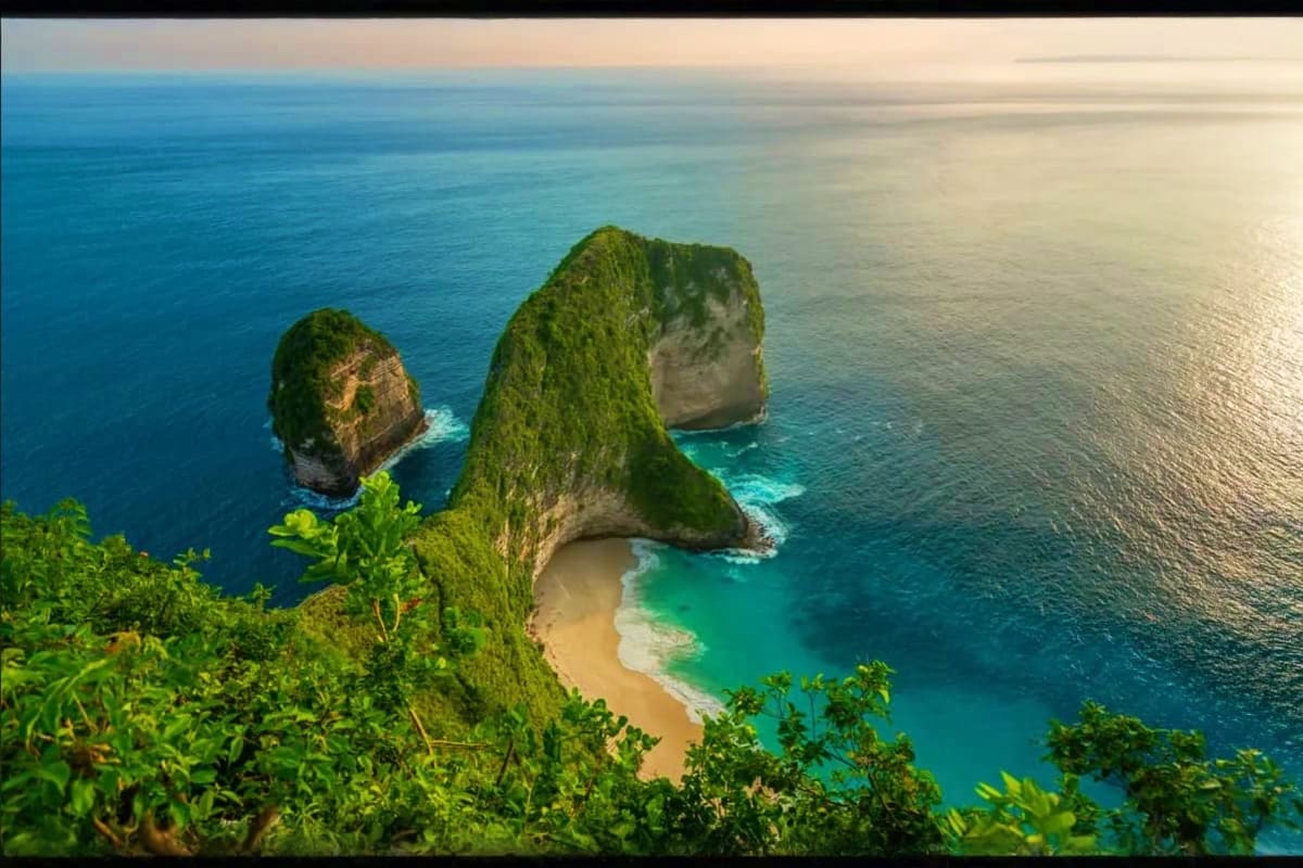 Aerial view of Kelingking Beach in Bali with turquoise ocean, white sand cove, and lush green cliff formations surrounded by