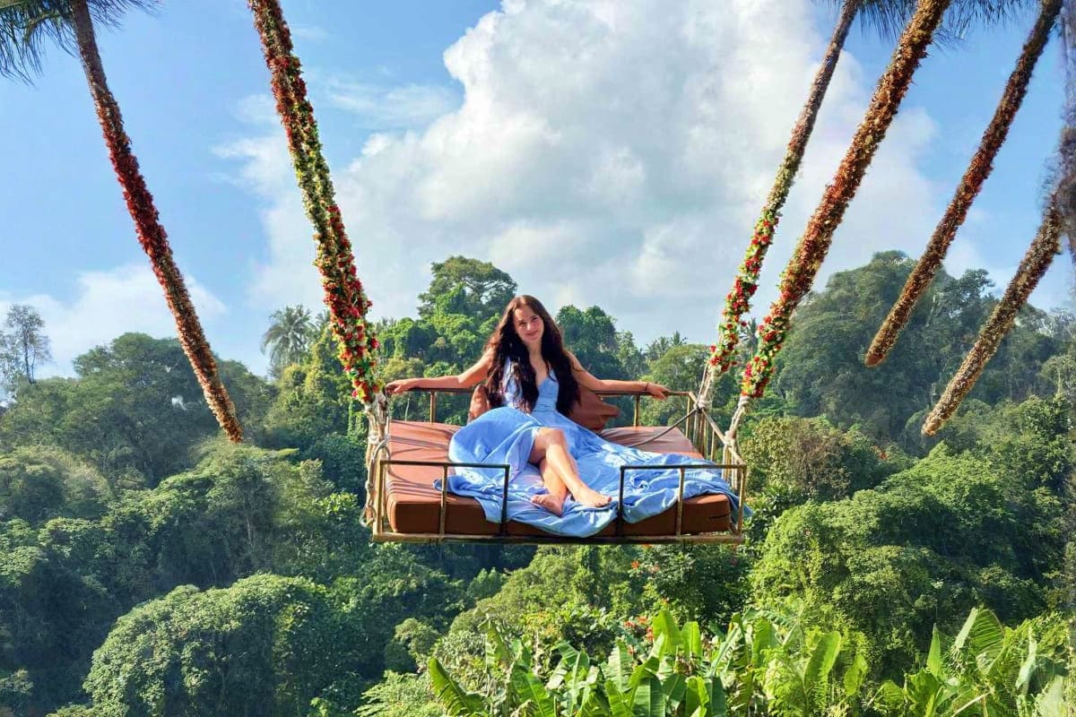 Woman sitting on decorative swing suspended over tropical jungle canopy in Bali, surrounded by flowering palm tree trunks