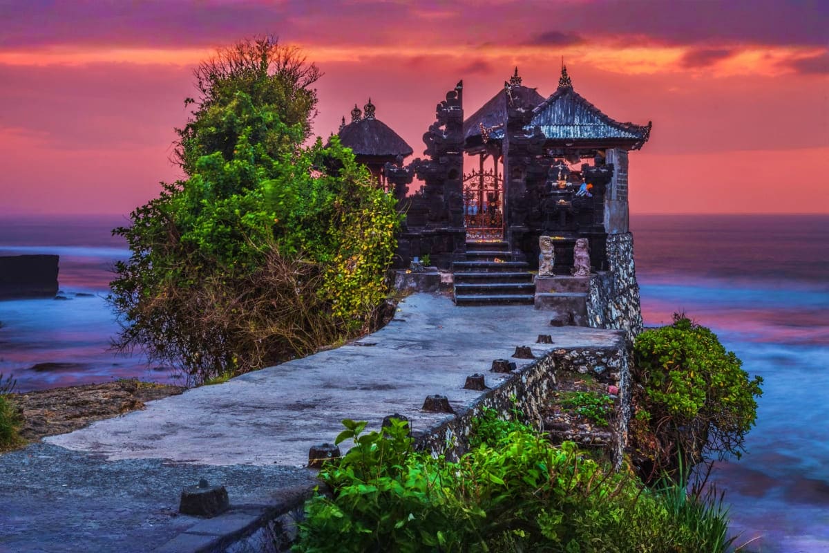 Ancient Balinese temple at sunset with ornate peaked roofs, stone pathway, lush green vegetation, and colorful purple-pink sk