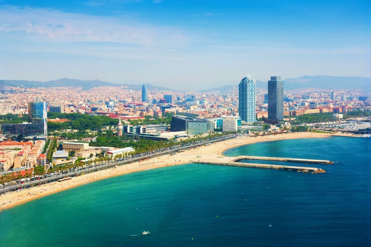 Aerial view of Barcelona's Barceloneta Beach with turquoise Mediterranean waters, sandy shoreline, palm trees, and city skyli