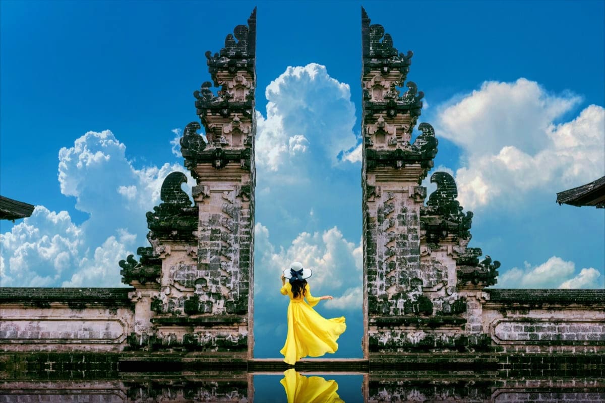 Family Paradise In Bali Woman in yellow dress standing between ornate stone temple gates in Bali with blue sky and clouds reflected in water below