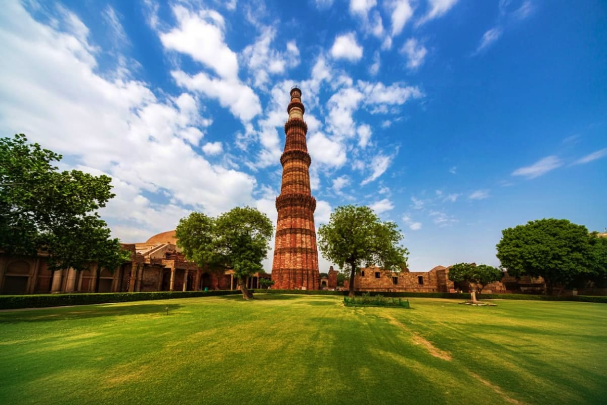 Qutb Minar red brick tower standing tall in green courtyard with historic ruins and blue sky with white clouds in Delhi