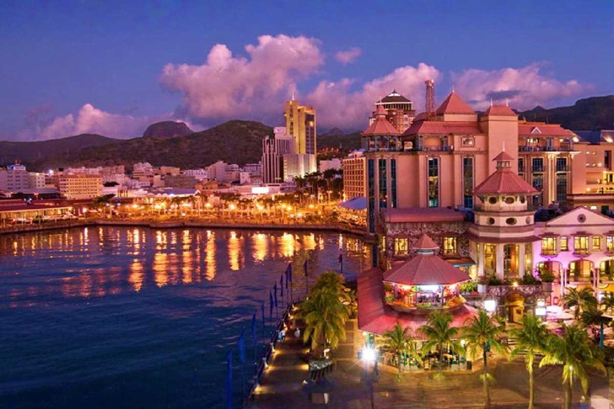 Waterfront promenade in Port Louis, Mauritius at dusk with illuminated colonial architecture, palm trees, and mountains in ba