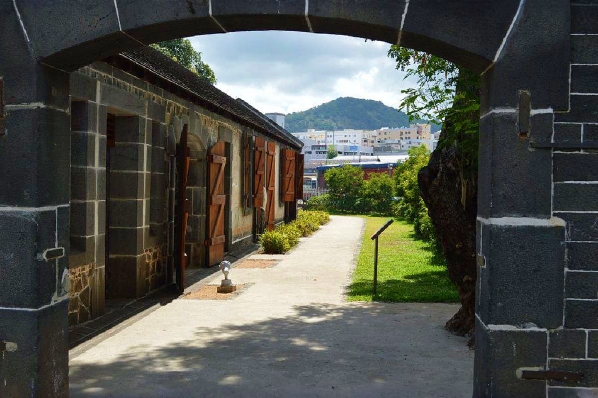 Historic stone fortification with wooden doors and grassy grounds, mountain and modern buildings visible in background, Mauri
