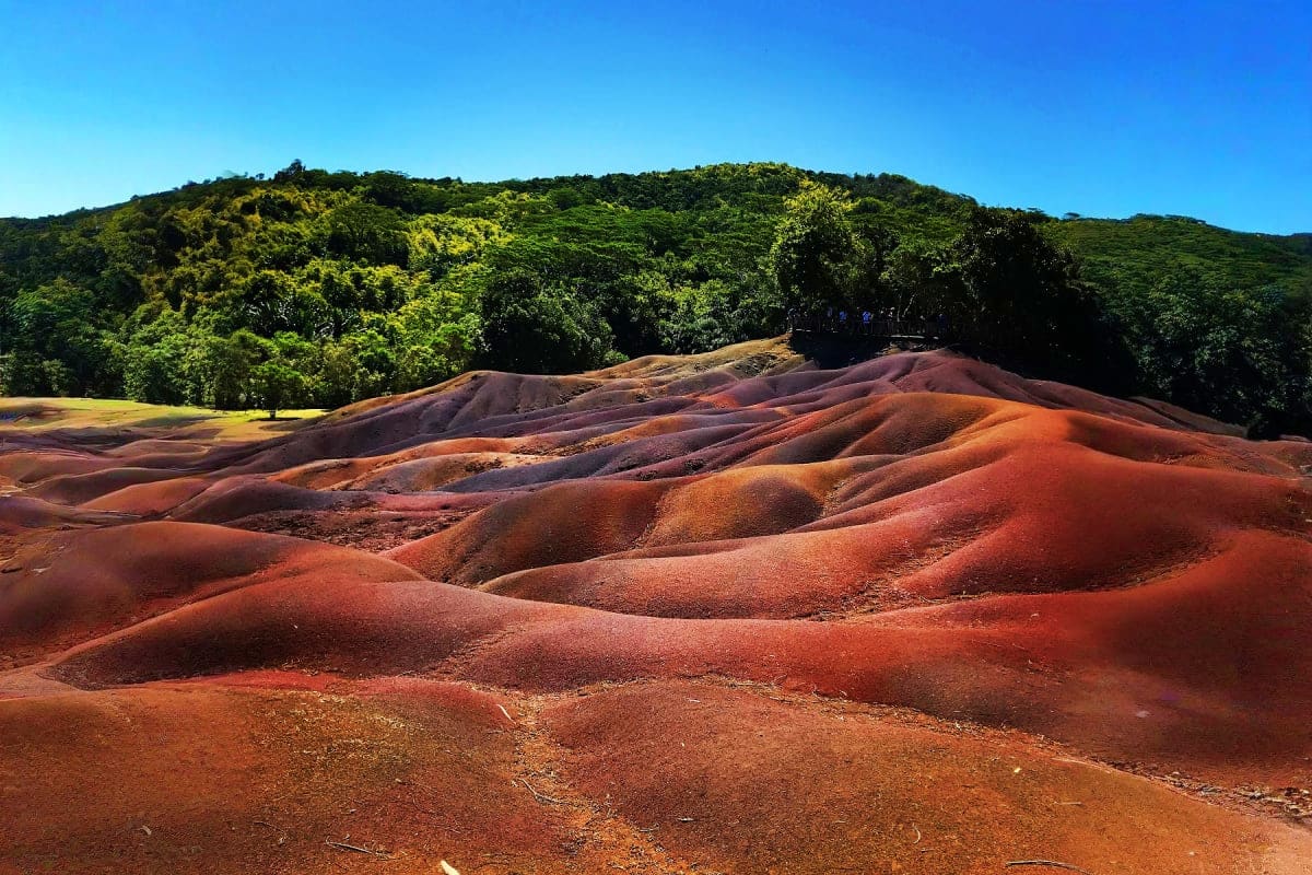 Colorful rolling hills of red, orange, and brown volcanic soil with lush green vegetation in the background under clear blue
