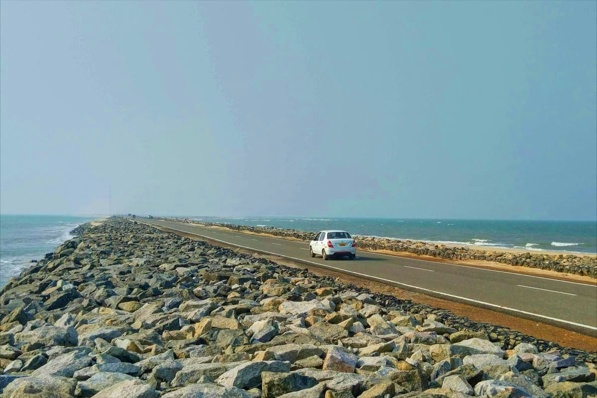 Temple and Coastline Highlights Madurai to Trivandrum White car driving on coastal road alongside rocky breakwater with Arabian Sea and sandy beach beyond