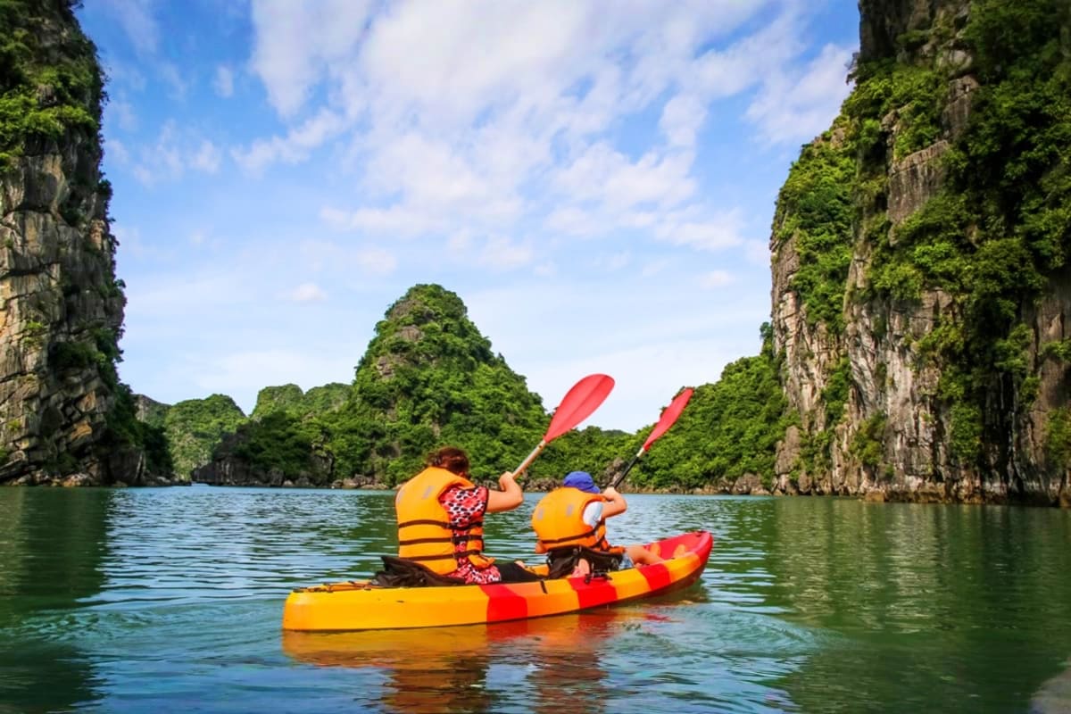 Two people kayaking in a yellow and red kayak with red paddles on turquoise water surrounded by limestone cliffs in Halong Ba